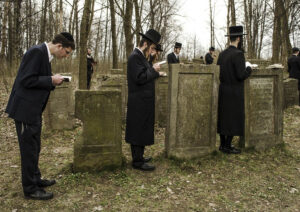 Grupa mężczyzn stoi pojedynczo przy nagrobkach na żydowskim cmentarzu. Modlą się odmawiając kadisz – modlitwę wyrażającą poddanie się woli Boga. W dłoniach trzymają książki, niekiedy opierając je o macewy. Kilku Żydów ubranych jest w długie chałaty, na głowach mają wysokie kapelusze z szerokimi rondami. Pozostali mają czapki lub jarmułki. Cmentarz porastają liczne drzewa i krzewy.